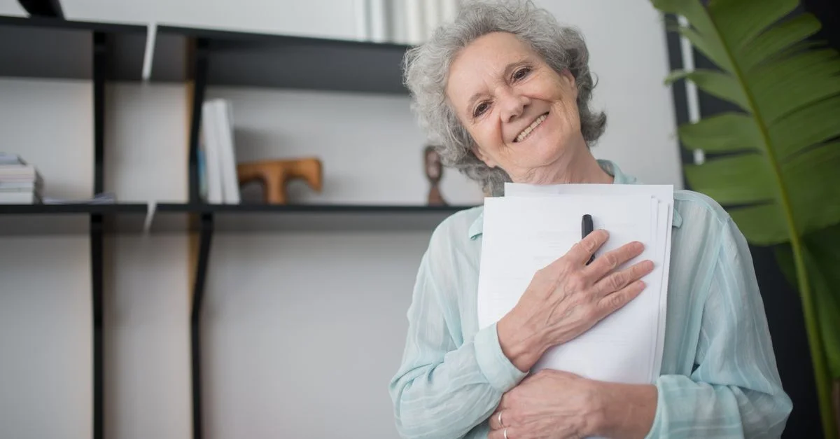 Femme senior souriante tenant des documents à l'intérieur, montrant bonheur et contentement.